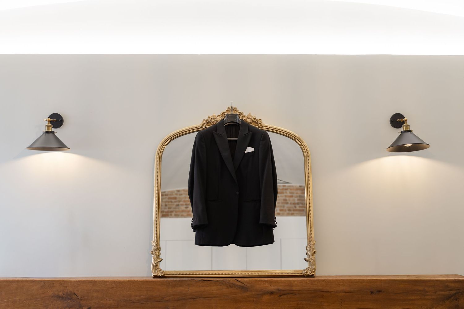 Black tuxedo jacket hanging on ornate gold mirror frame between two vintage wall sconces.