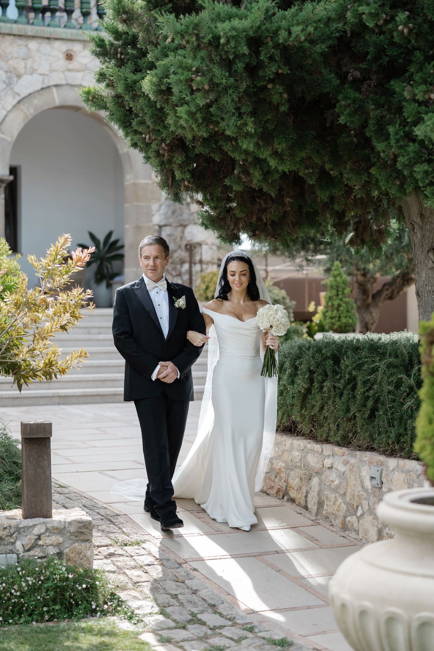 Bride in white off-shoulder gown walks with escort through garden venue.
