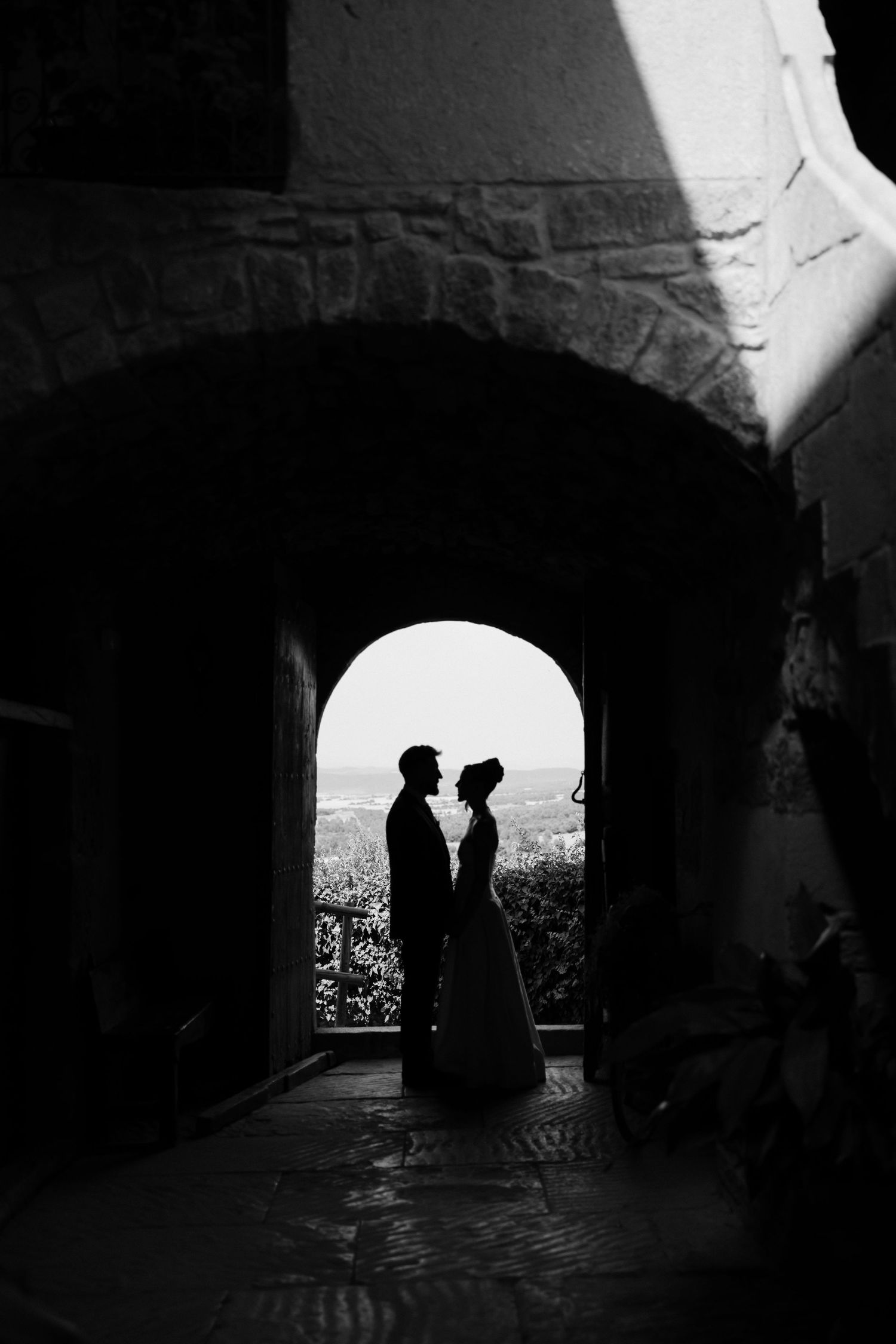 Silhouette of bride and groom facing each other framed by stone archway with scenic landscape behind them.