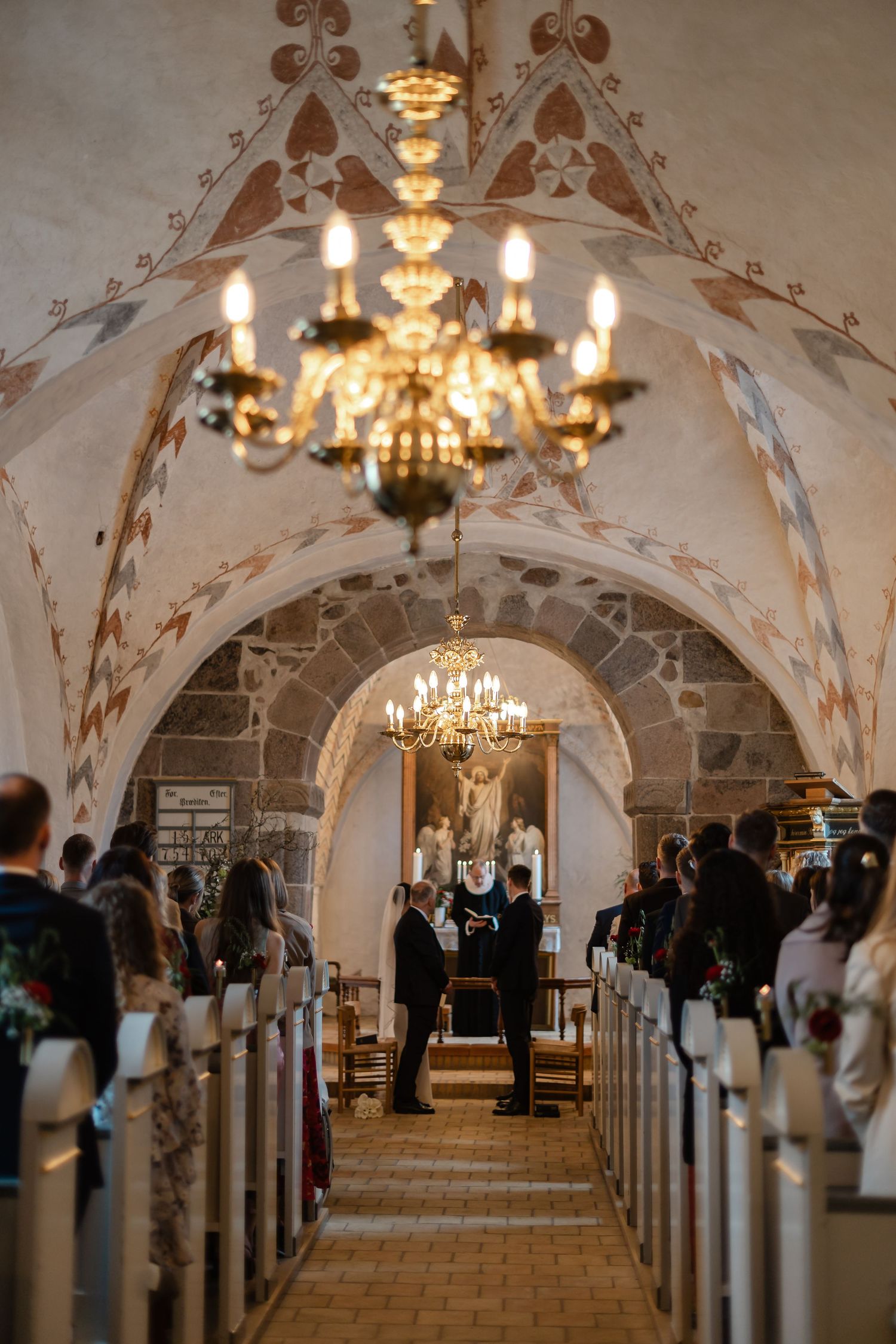 Wedding ceremony inside a historic stone church with vaulted ceilings, chandeliers, and painted arches as guests watch.