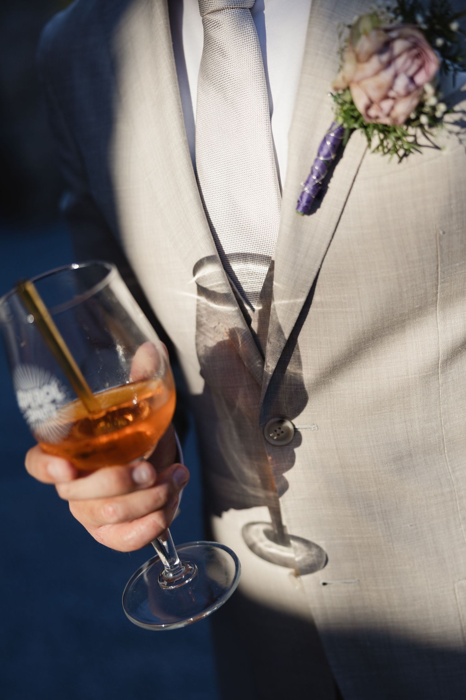 Groom in beige suit holding an aperol spritz cocktail with floral boutonniere at wedding.