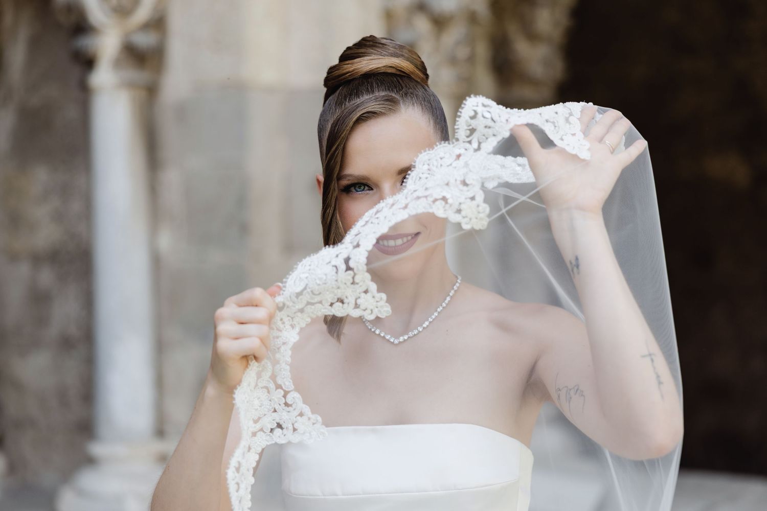 Beautiful bride holding lace veil over her face with a soft smile in black and white.