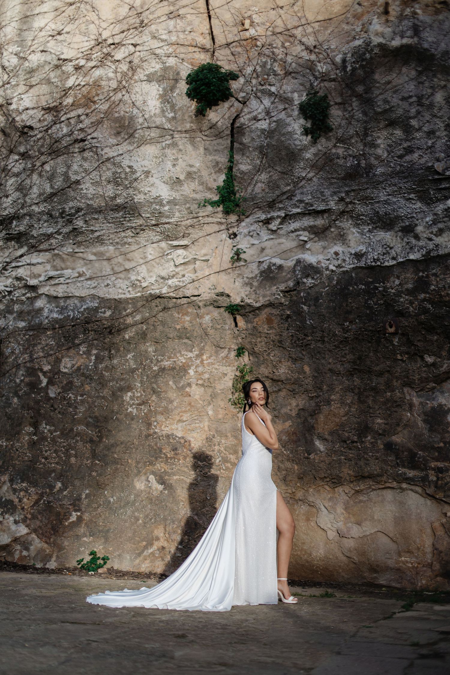 Bride in a flowing white gown with a long train standing before a dramatic textured stone wall.