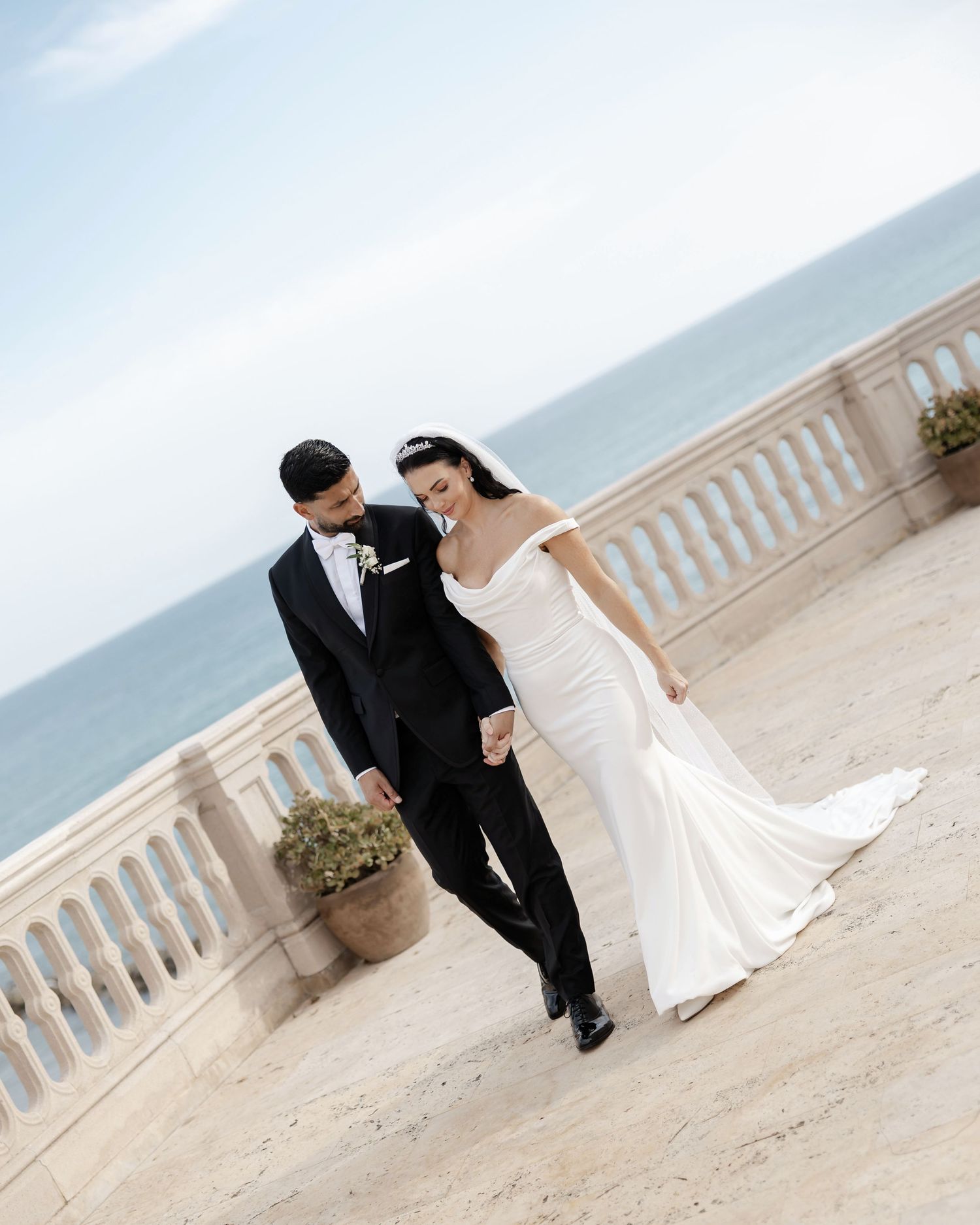 Bride and groom walking hand in hand on a seaside terrace, bride in off-shoulder mermaid gown, groom in black tuxedo.