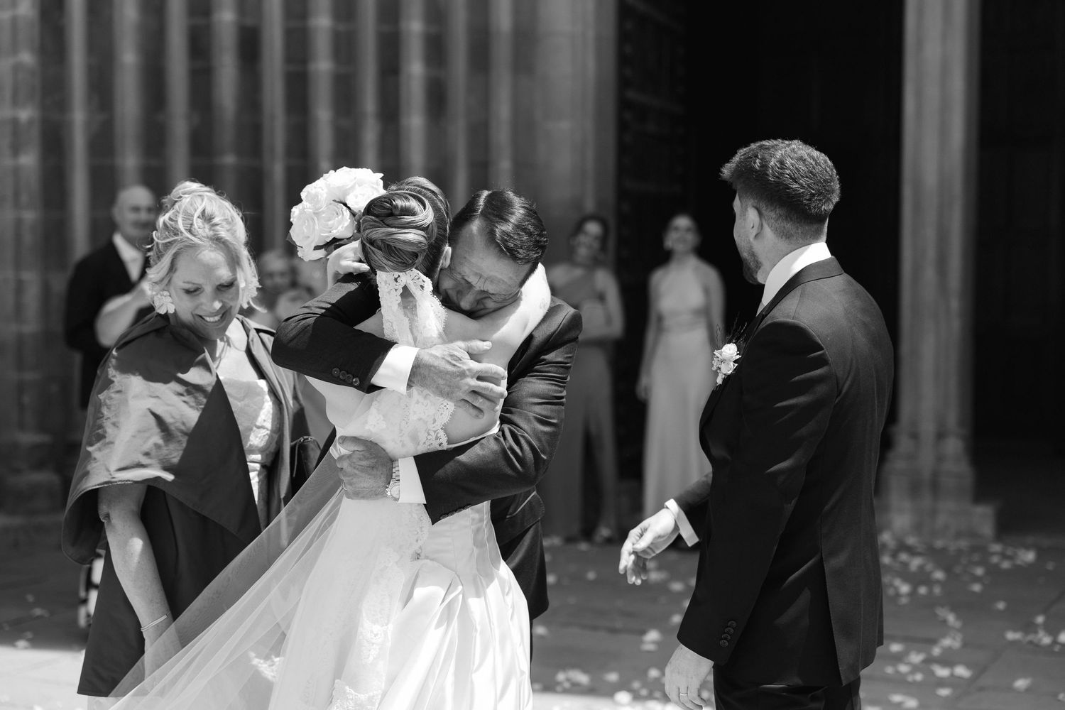 Black and white wedding photo of bride and groom sharing a kiss at church exit with guests celebrating nearby.