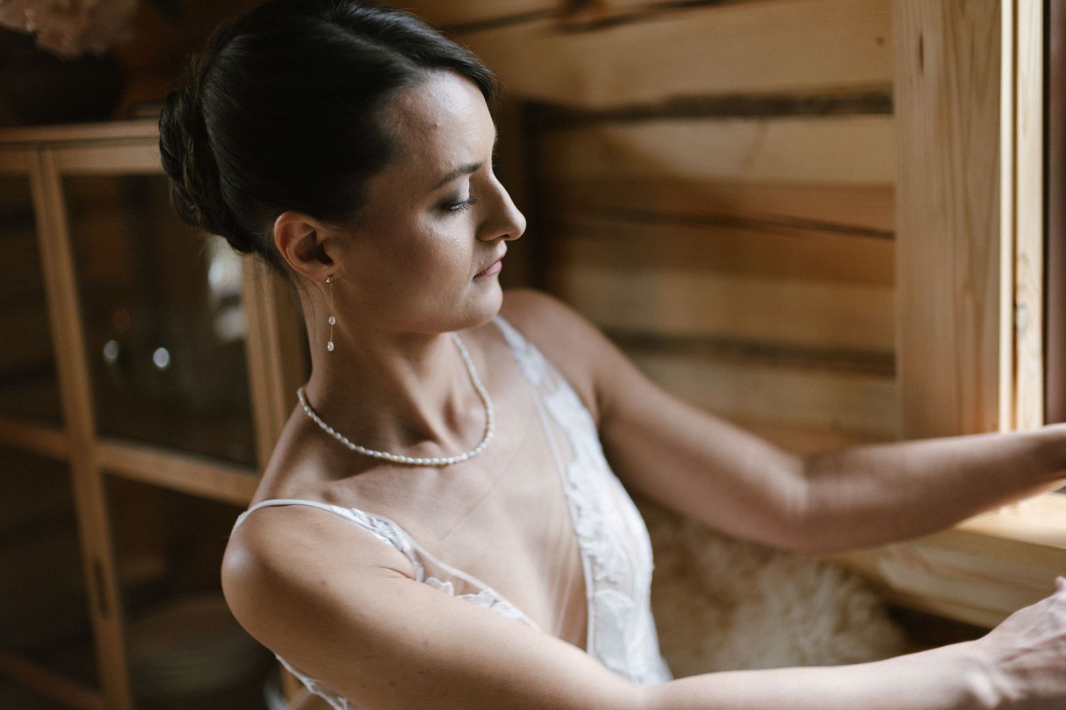 Beautiful bride in lace dress wearing pearl necklace and earrings sitting in a wooden sauna.