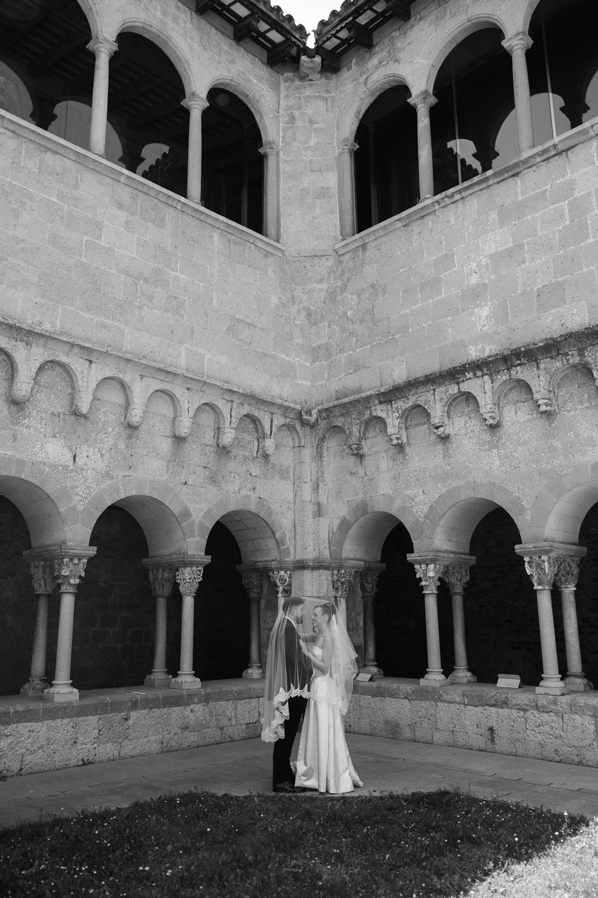 Black and white photo of a bride and groom embracing in a historic stone cloister with arched columns.