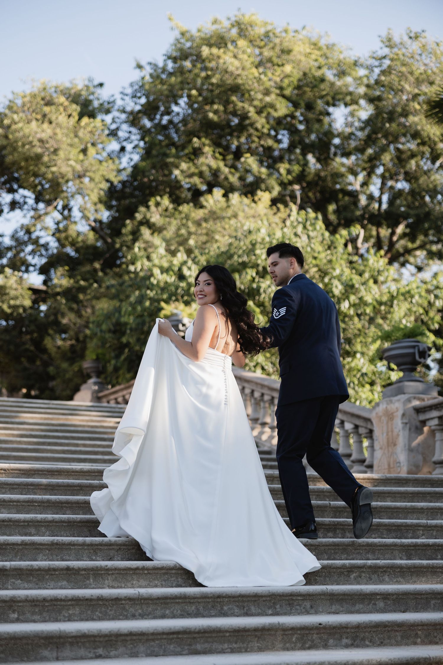Bride and groom walking up stone steps outdoors, bride in white gown, groom in navy suit, lush green trees behind.