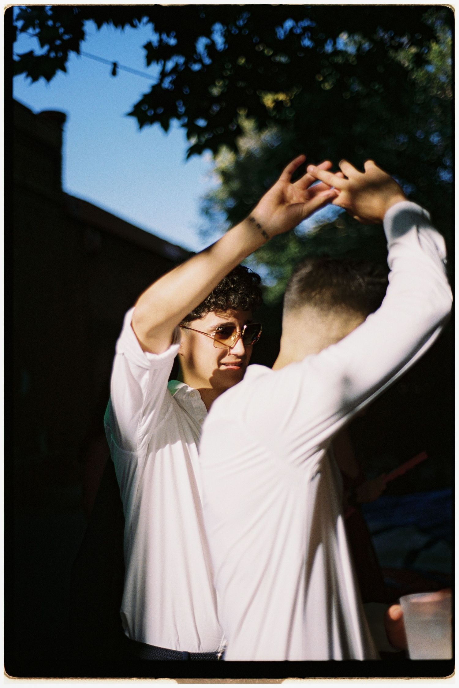 Two men in white shirts dance together outdoors under string lights and trees on a sunny day.