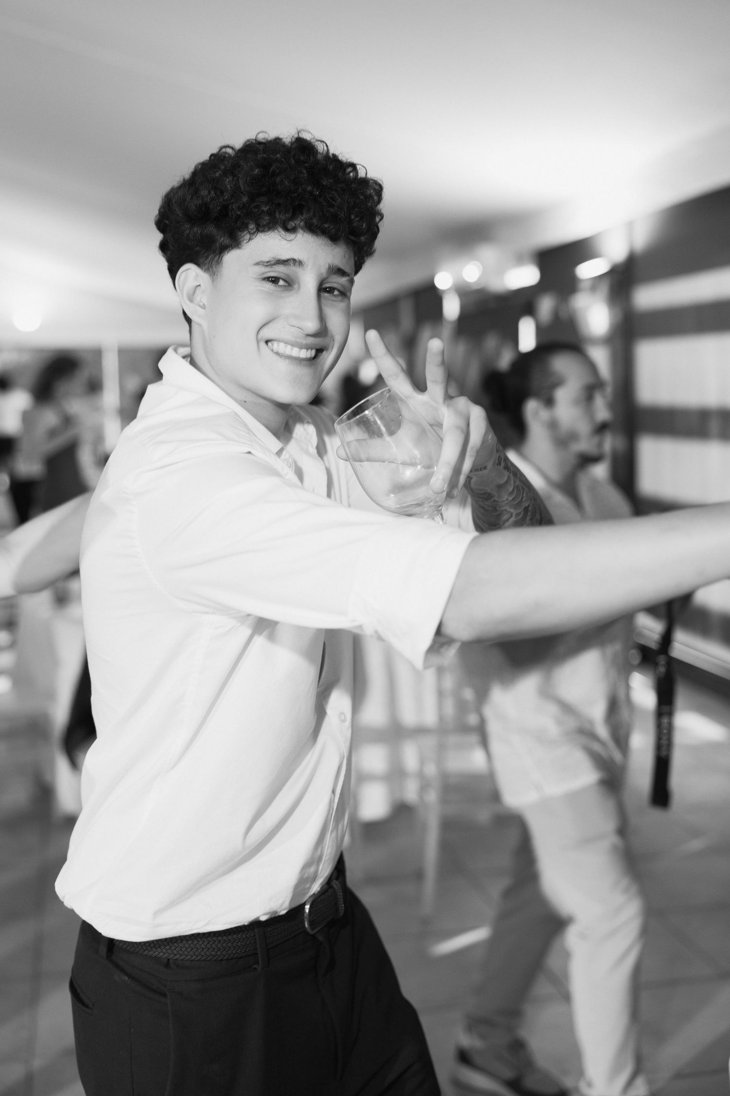 Young man with curly hair in white shirt smiling and dancing at a wedding reception in black and white.