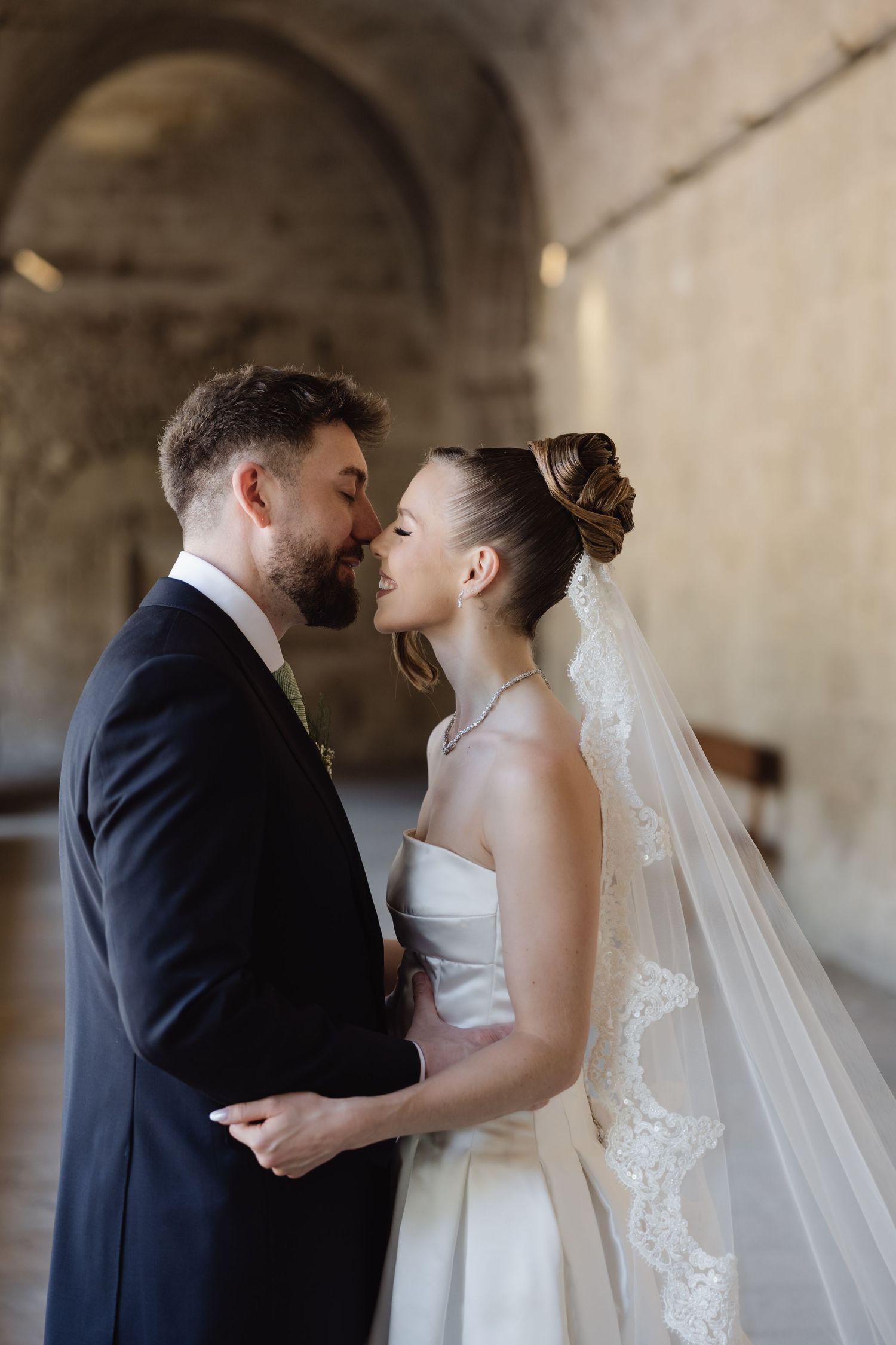 Bride and groom share intimate moment in historic stone archway, bride wearing strapless gown and lace veil.