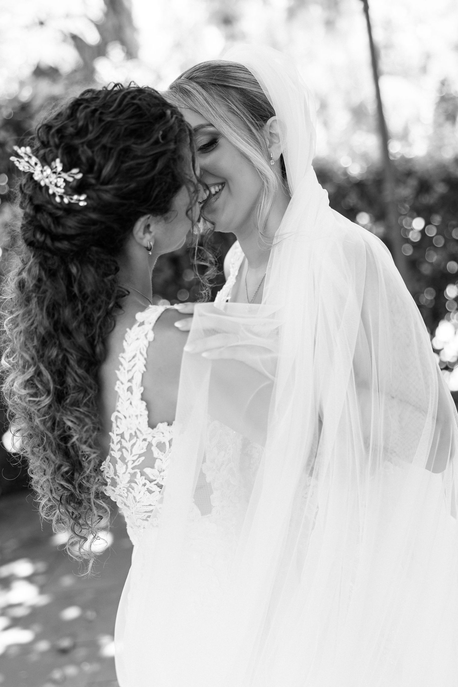 Two brides in white lace gowns share an intimate moment touching foreheads in a black and white photo.