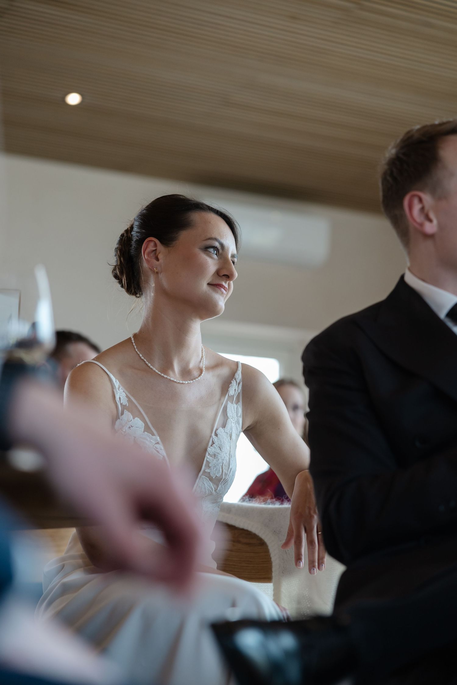 Elegant bride in white gown with pearl necklace smiles warmly during wedding ceremony while seated beside groom.