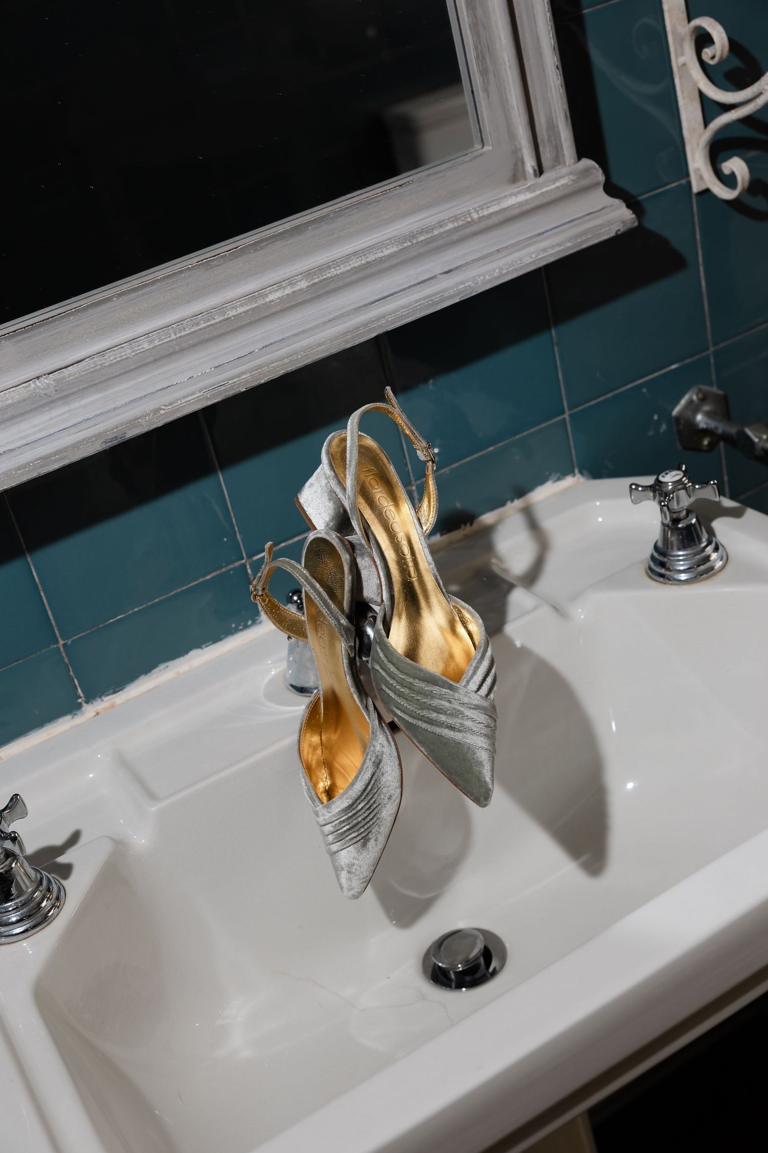Silver slingback heels resting on a white bathroom sink with teal tiles and vintage faucets.