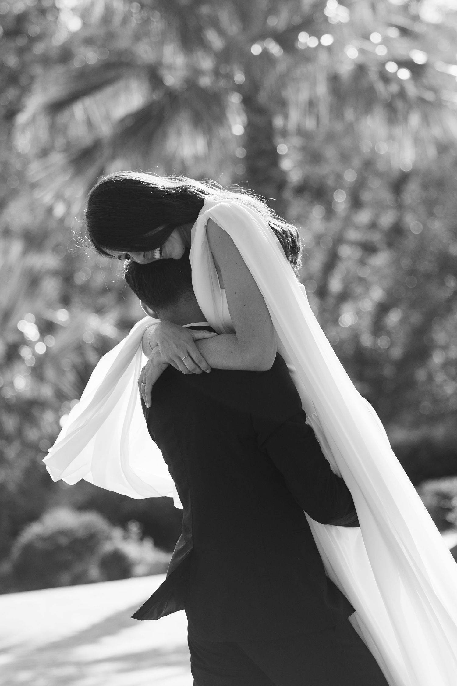 Black and white photo of bride and groom sharing an emotional embrace outdoors with palm trees in background.