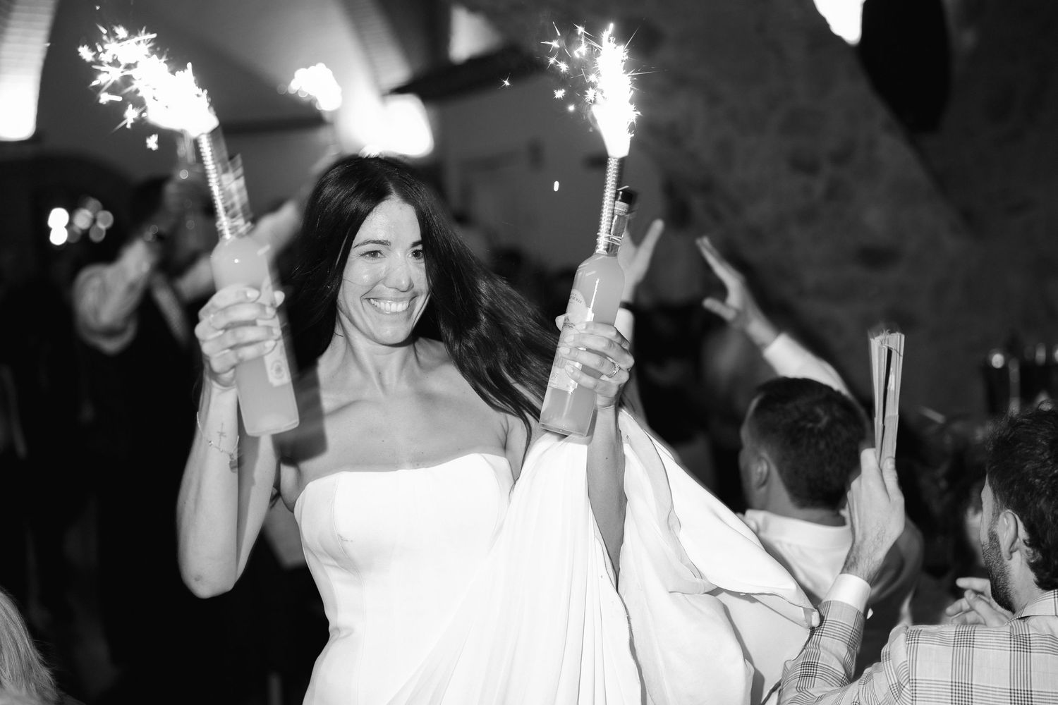 Smiling bride in white dress holding sparklers at wedding reception in black and white photo.