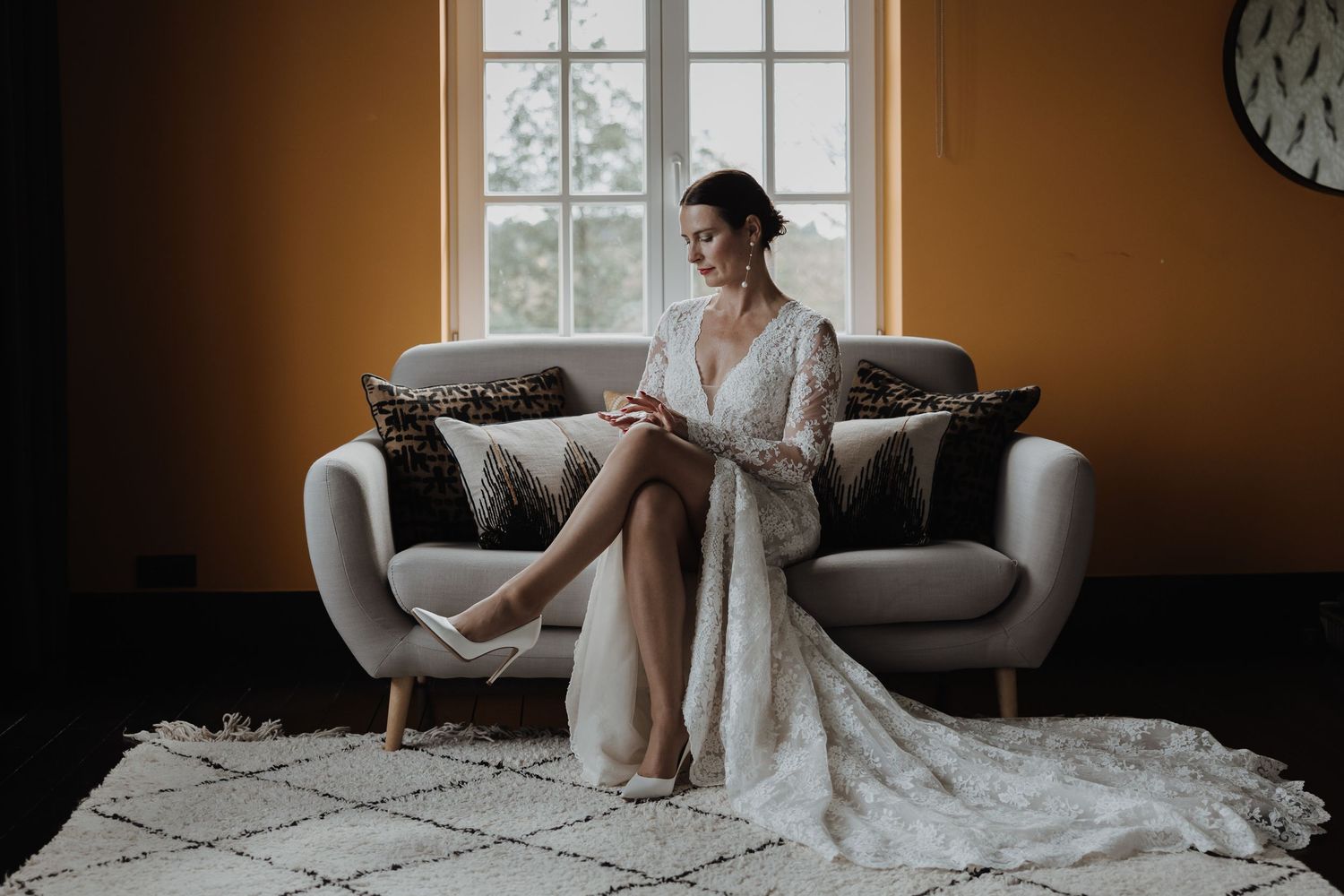 Bride in long-sleeve lace wedding gown sitting on a sofa in a golden room, adjusting her heel.