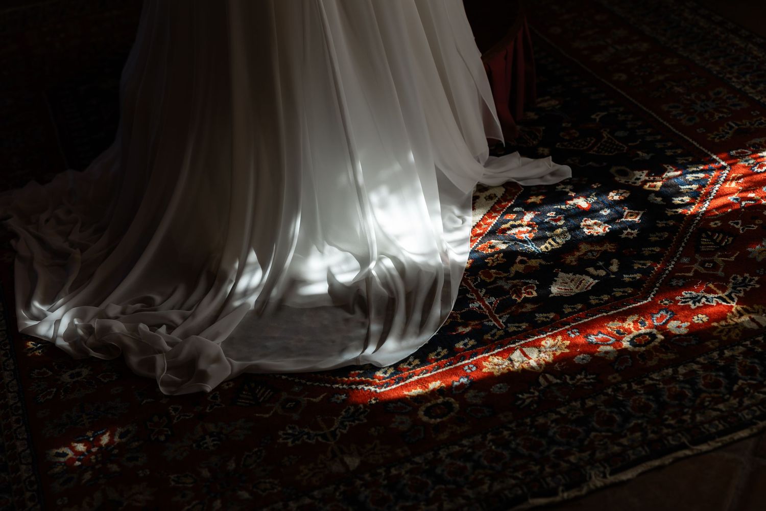 Close-up of white wedding gown hem glowing in dramatic light on ornate patterned rug.