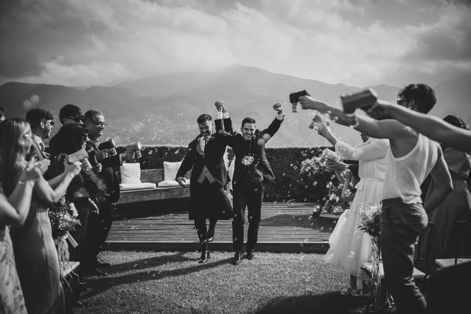 Black and white photo of two grooms celebrating joyfully with wedding guests outdoors.