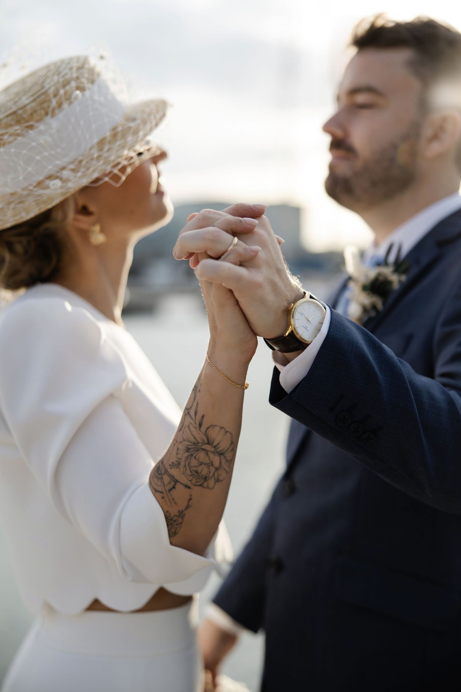 Bride with floral tattoo and straw hat holds hands with groom in navy suit at outdoor wedding.