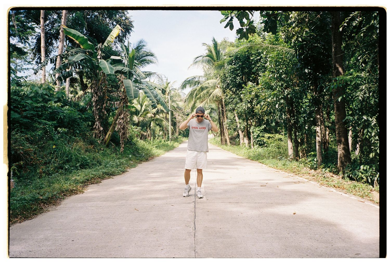 A man stands on a tropical road lined with palm trees in a sunny outdoor setting.