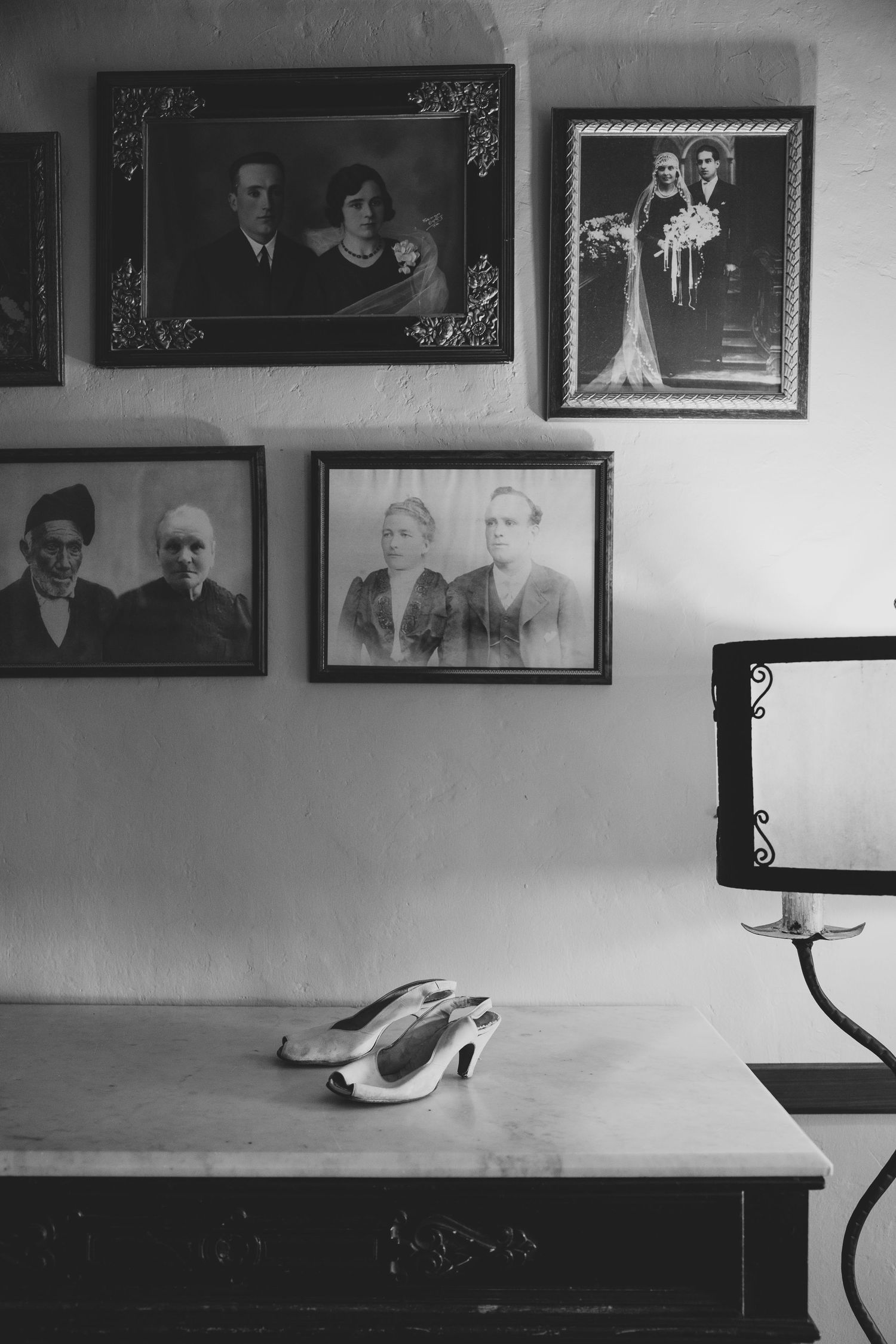 Black and white photo of vintage family portraits on wall above dresser with bridal shoes.