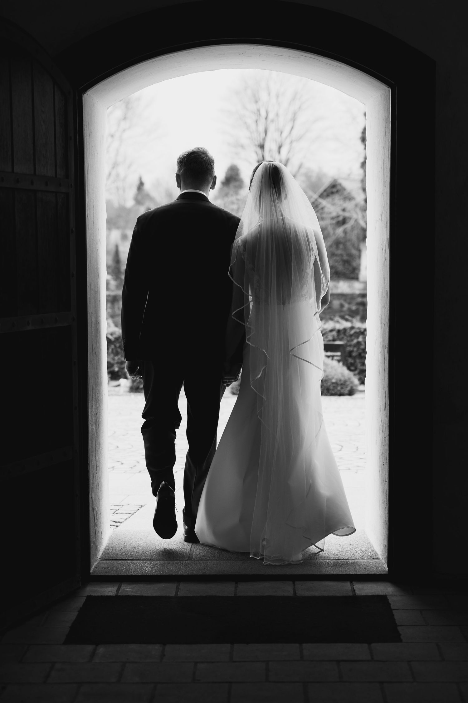 Black and white silhouette of bride and groom walking through arched church doorway after wedding ceremony.