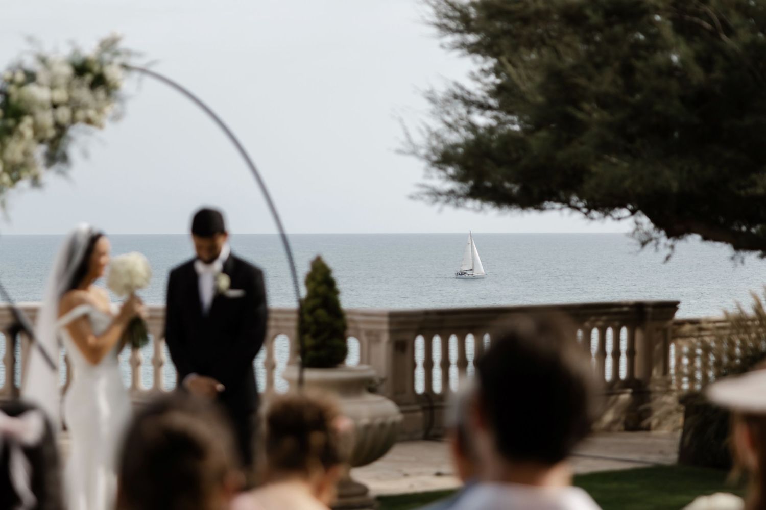 Outdoor wedding ceremony with floral arch framing couple against stunning ocean sailboat view.