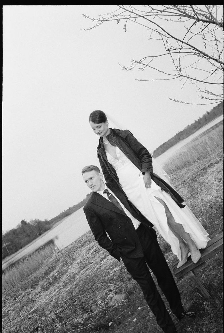 Black and white photo of a bride and groom posing near a lake with bare trees in the background.