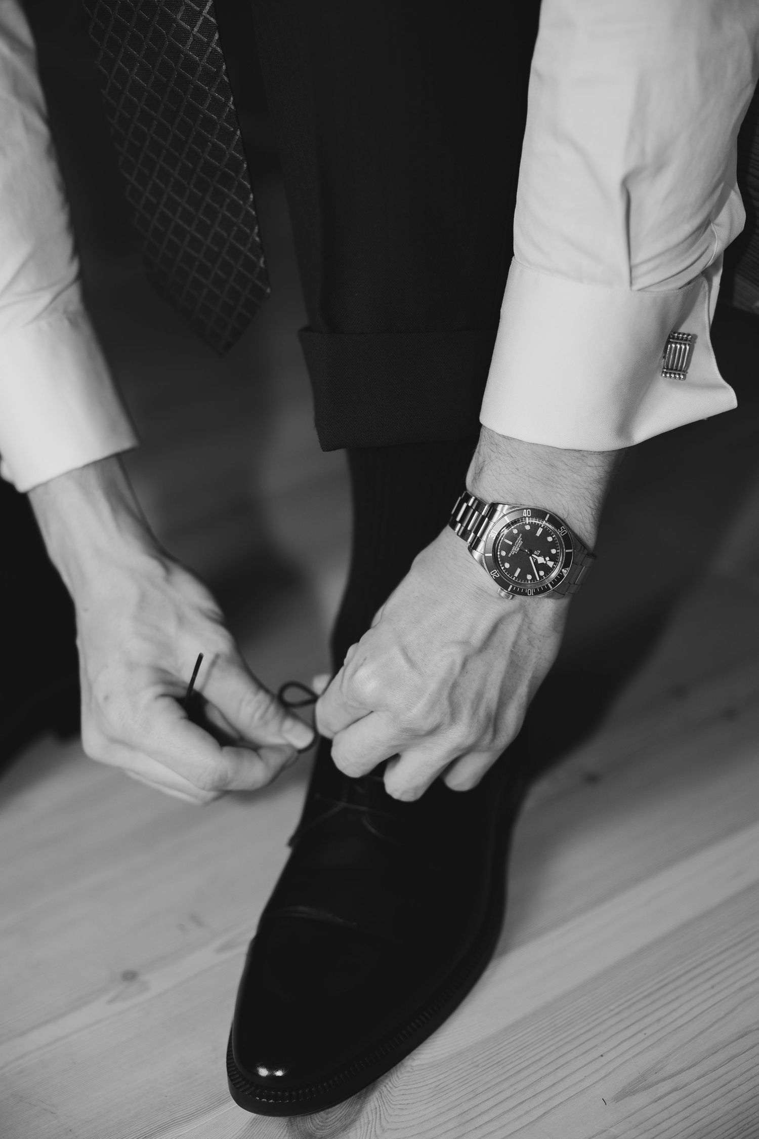 Groom tying his dress shoe laces wearing a suit, patterned tie, and silver watch on wedding day.