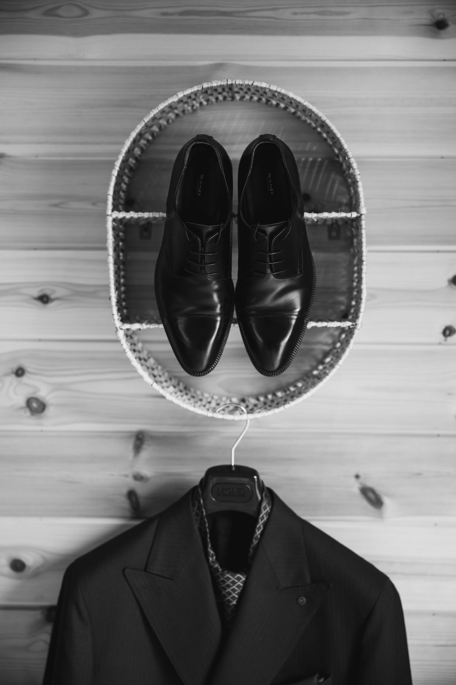 Black and white flat lay of mens dress shoes in oval tray above a suit jacket on hanger against wood wall.
