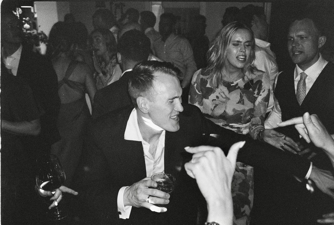 Black and white photo of people socializing at a formal party, man in suit holding drink laughing with blonde woman.