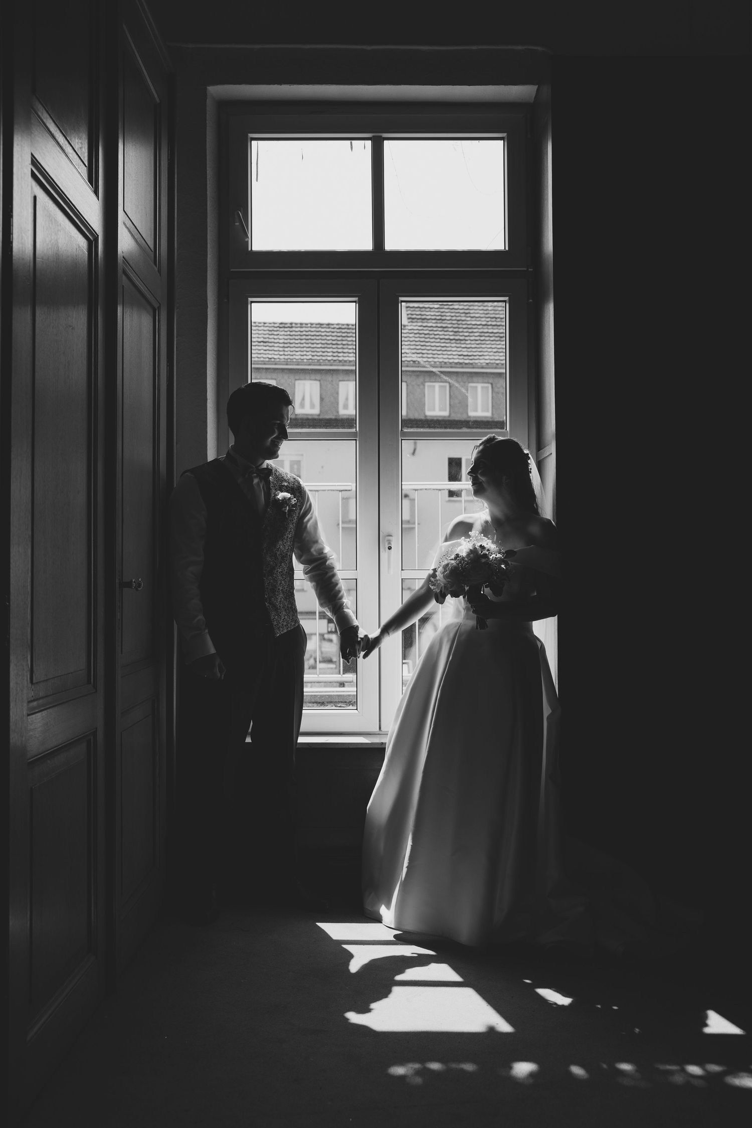 Black and white silhouette of bride and groom holding hands by a sunlit window.