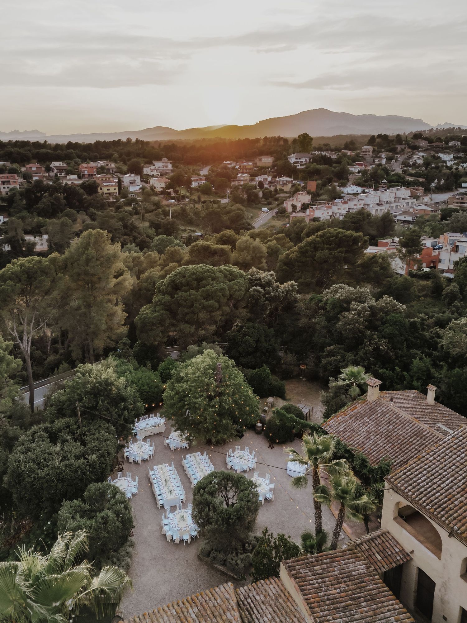 Aerial sunset view of elegant outdoor wedding reception venue surrounded by lush trees and mountains.