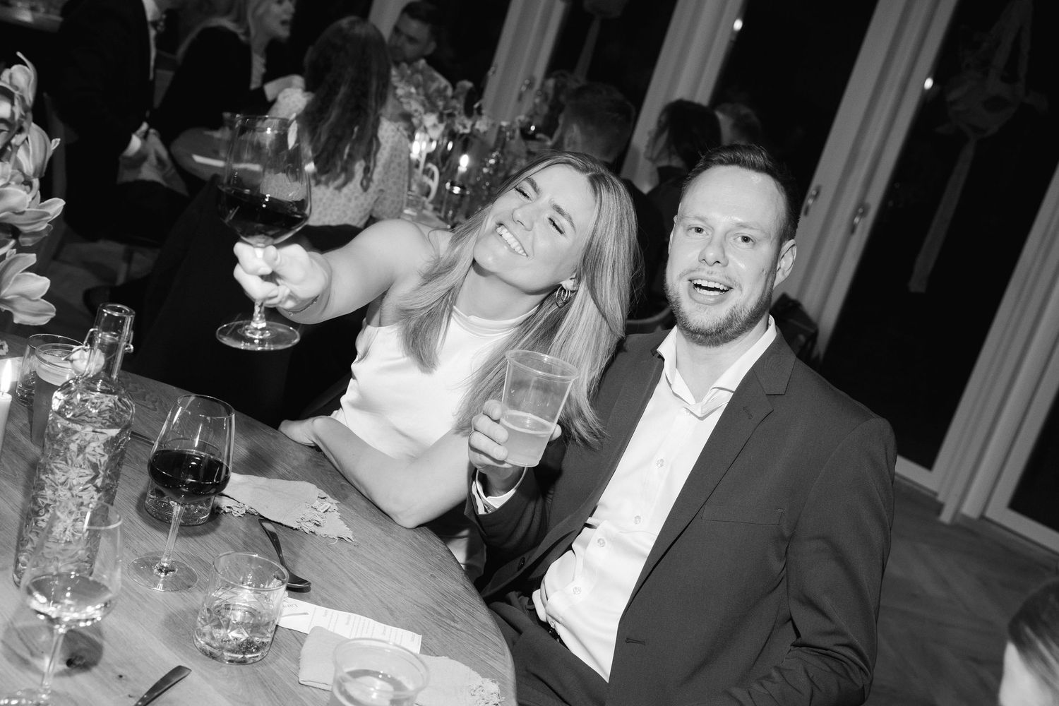 Black and white photo of a couple smiling and raising glasses of wine at a wedding reception dinner table.