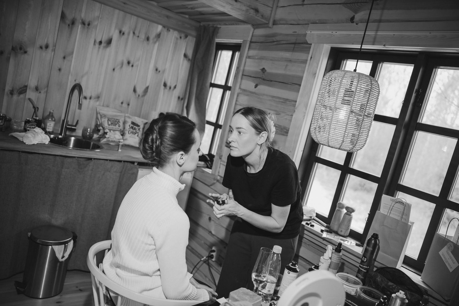 Black and white photo of a makeup artist applying makeup to a bride in a rustic wooden cabin interior.
