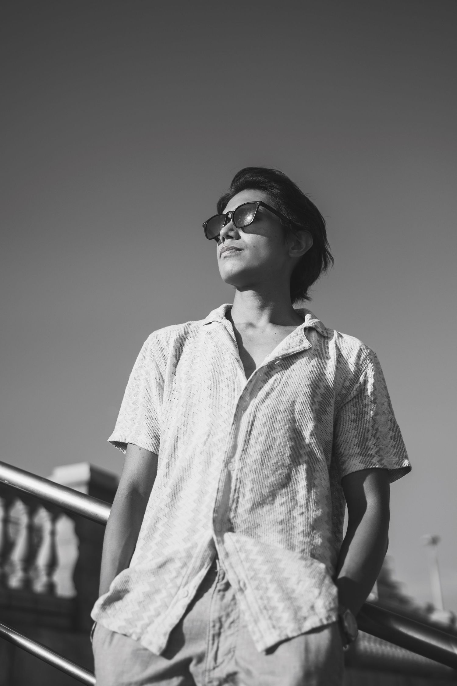Black and white portrait of a young man in sunglasses and short-sleeve shirt, standing outdoors against a clear sky.