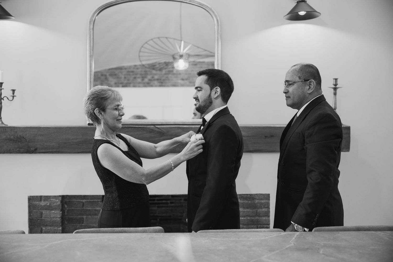 Black and white photo of woman adjusting grooms boutonniere while man stands nearby indoors.