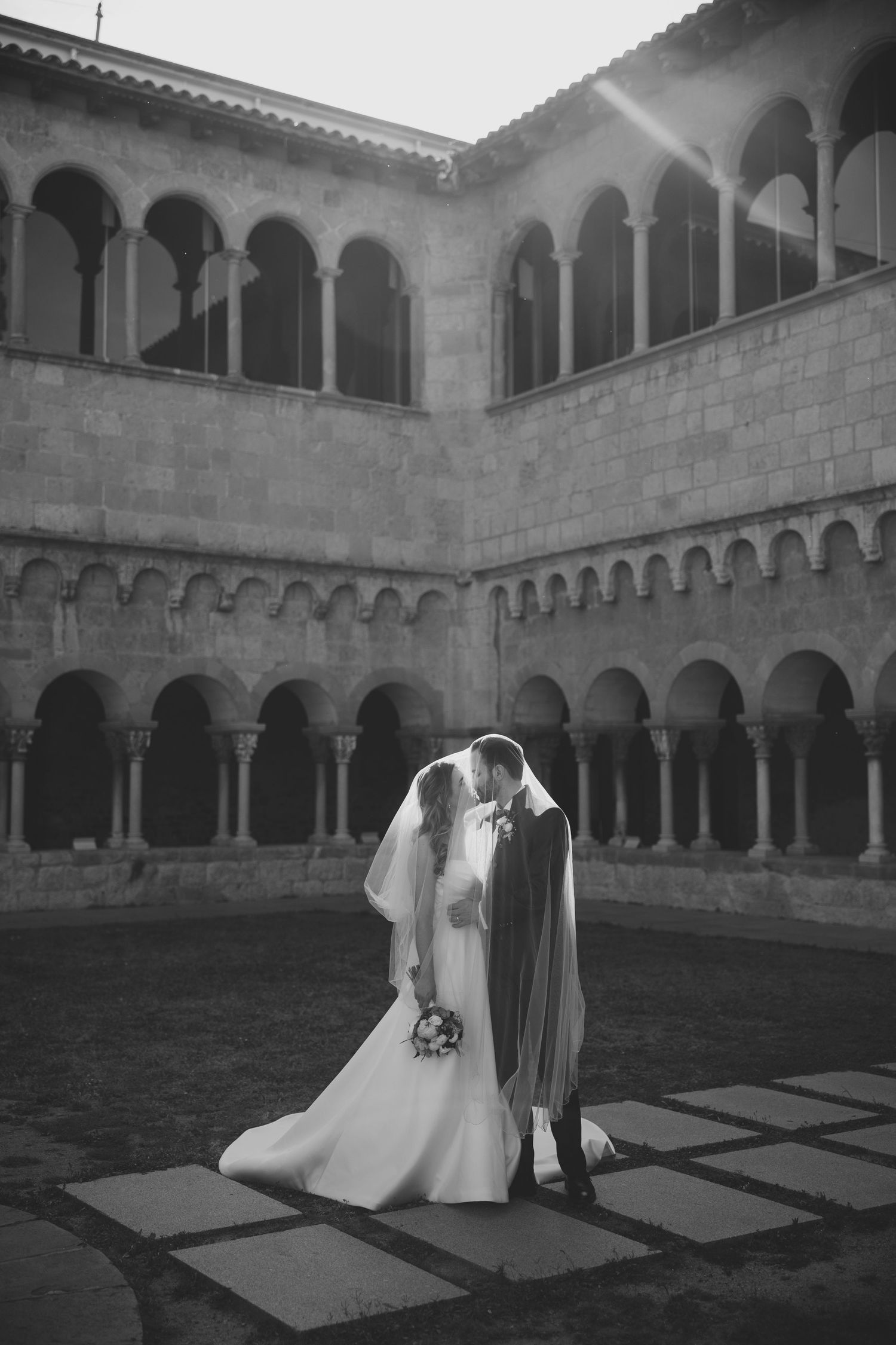 Black and white photo of bride and groom kissing in a historic stone courtyard.
