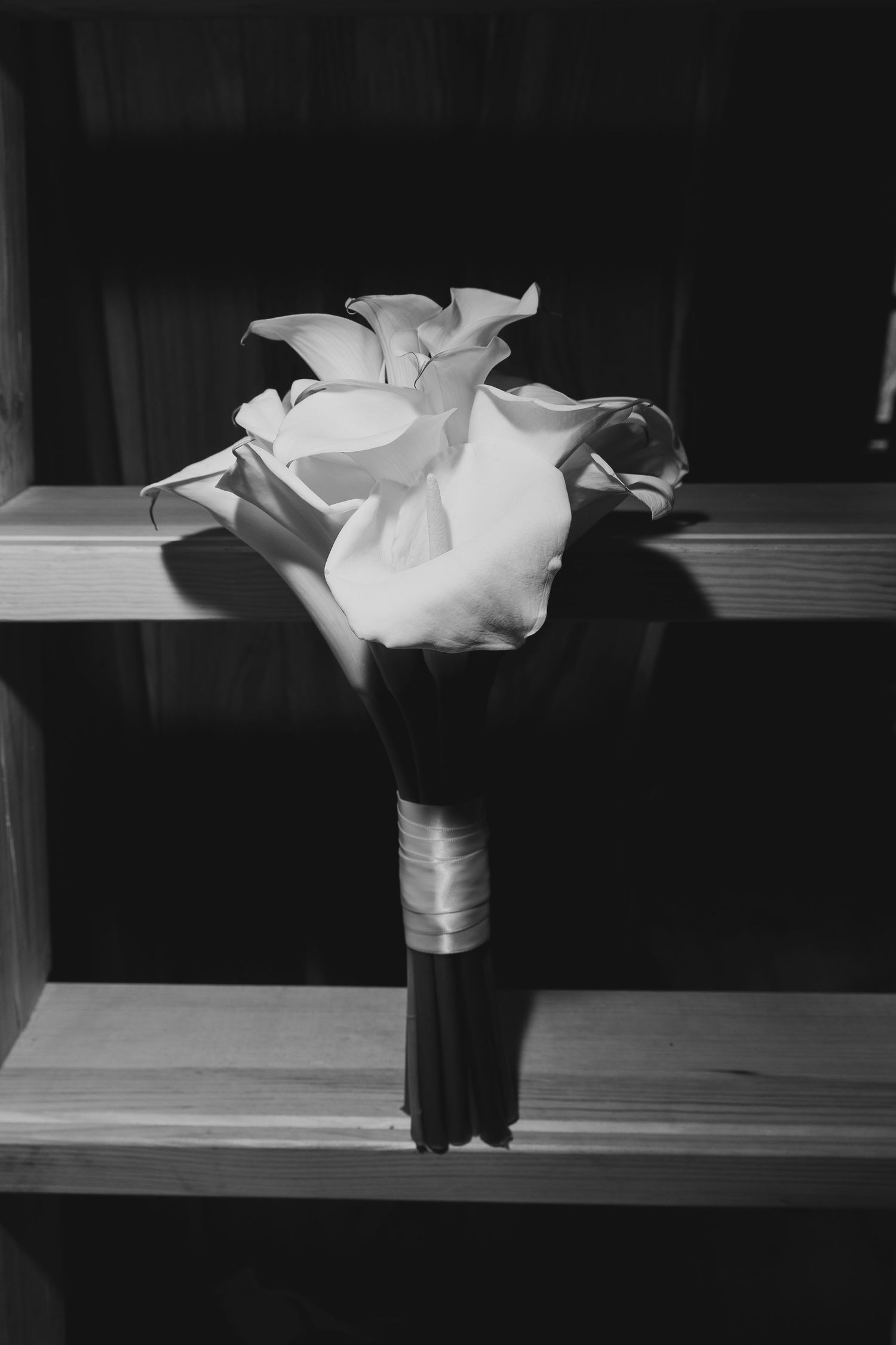 Black and white photo of elegant calla lily wedding bouquet resting on wooden steps, wrapped with ribbon.