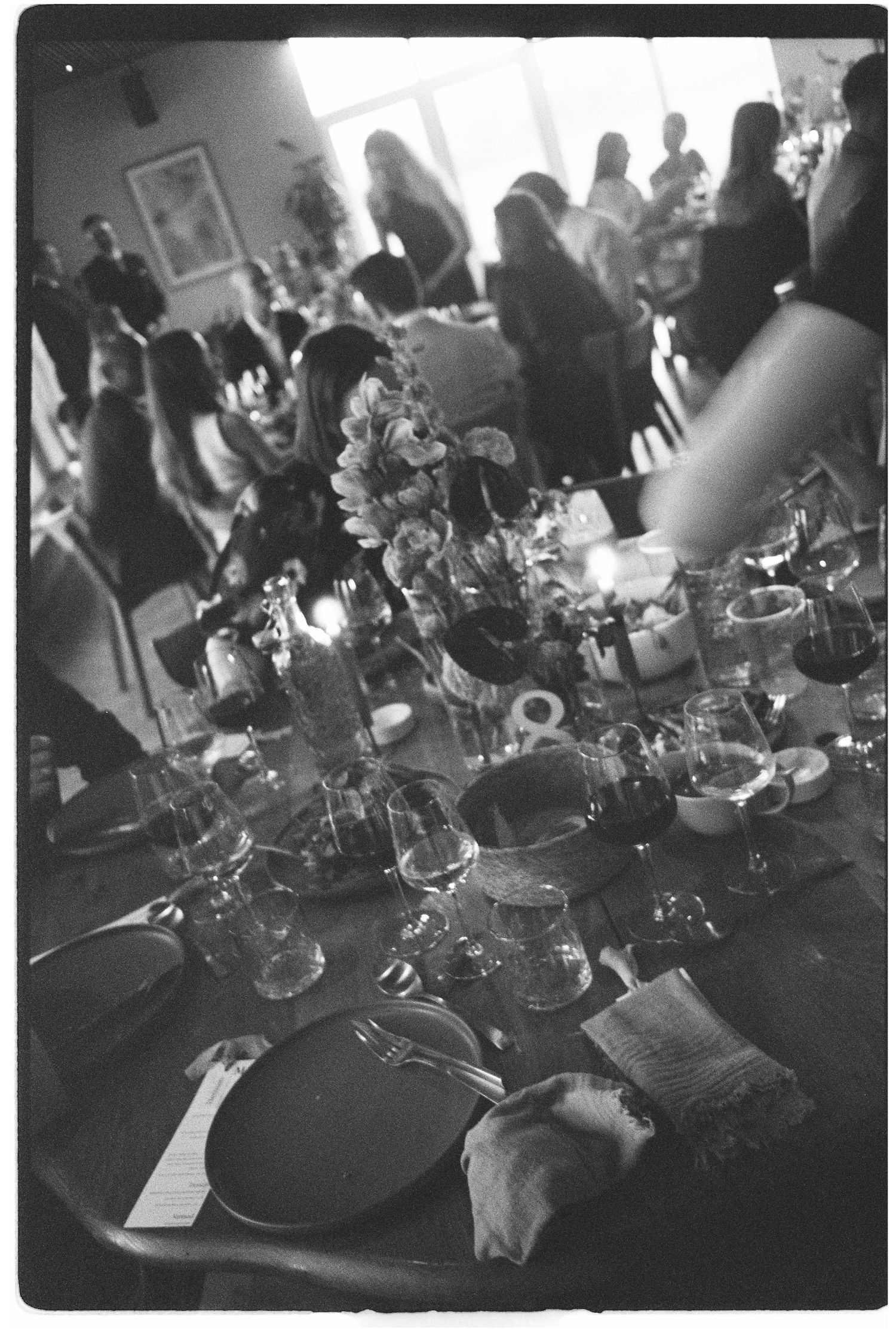 Black and white photo of an elegant dinner party table setting with floral centerpiece and guests mingling in background.