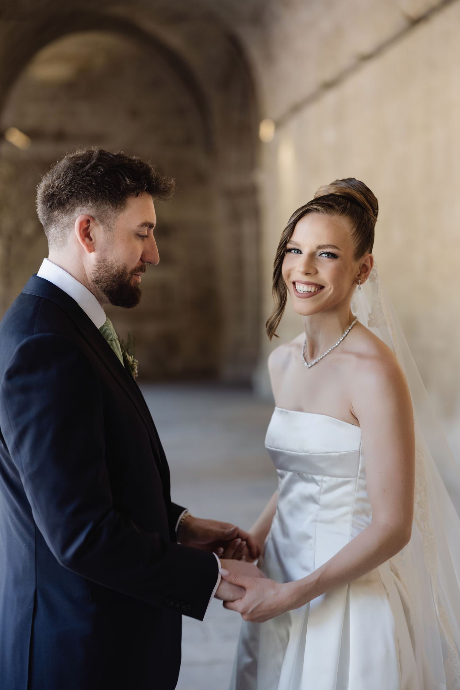 Smiling bride in white strapless gown holds hands with bearded groom in navy suit under stone archway.