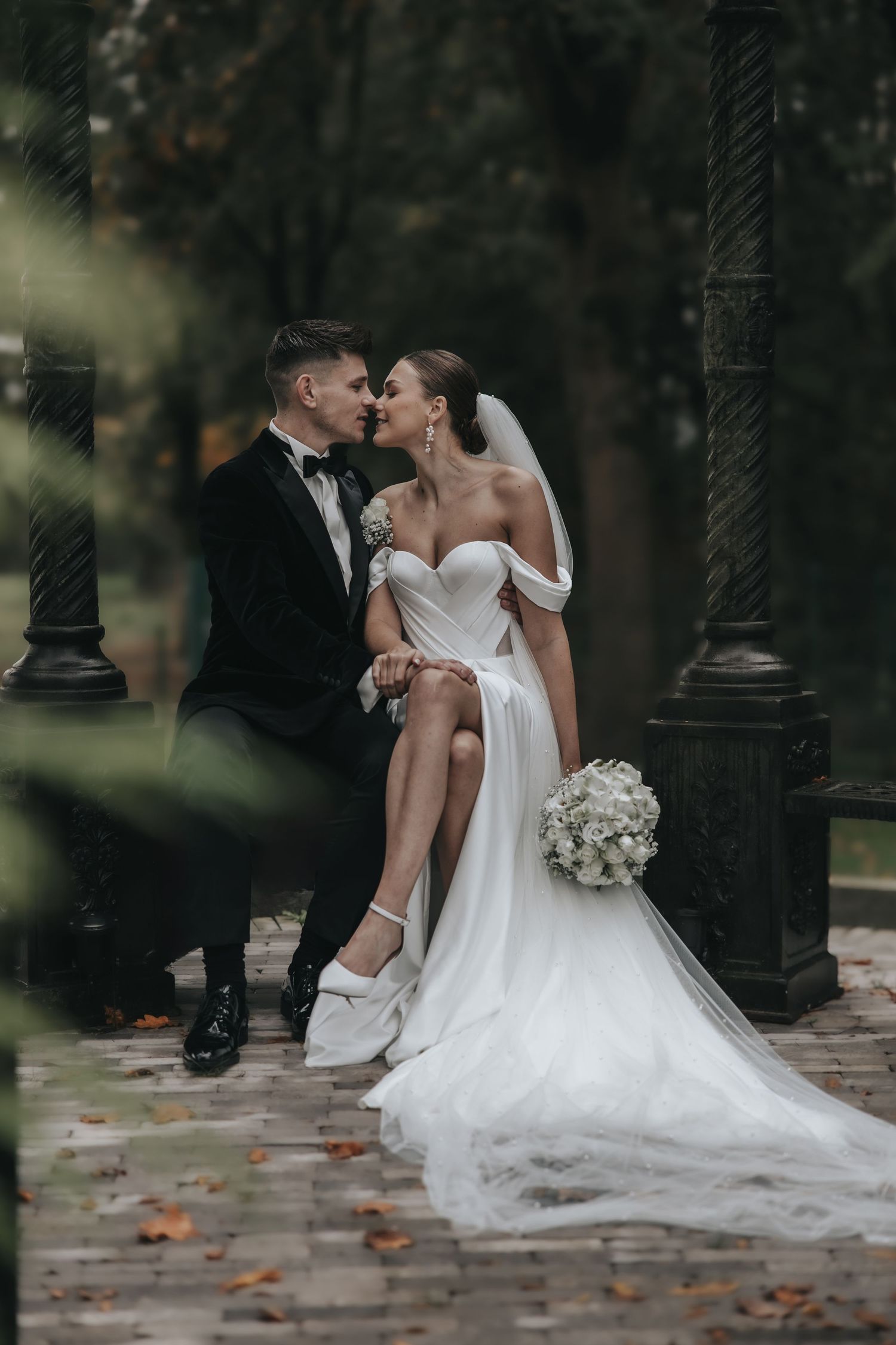 Bride and groom share a kiss outdoors in an autumn park, bride holding a white floral bouquet.