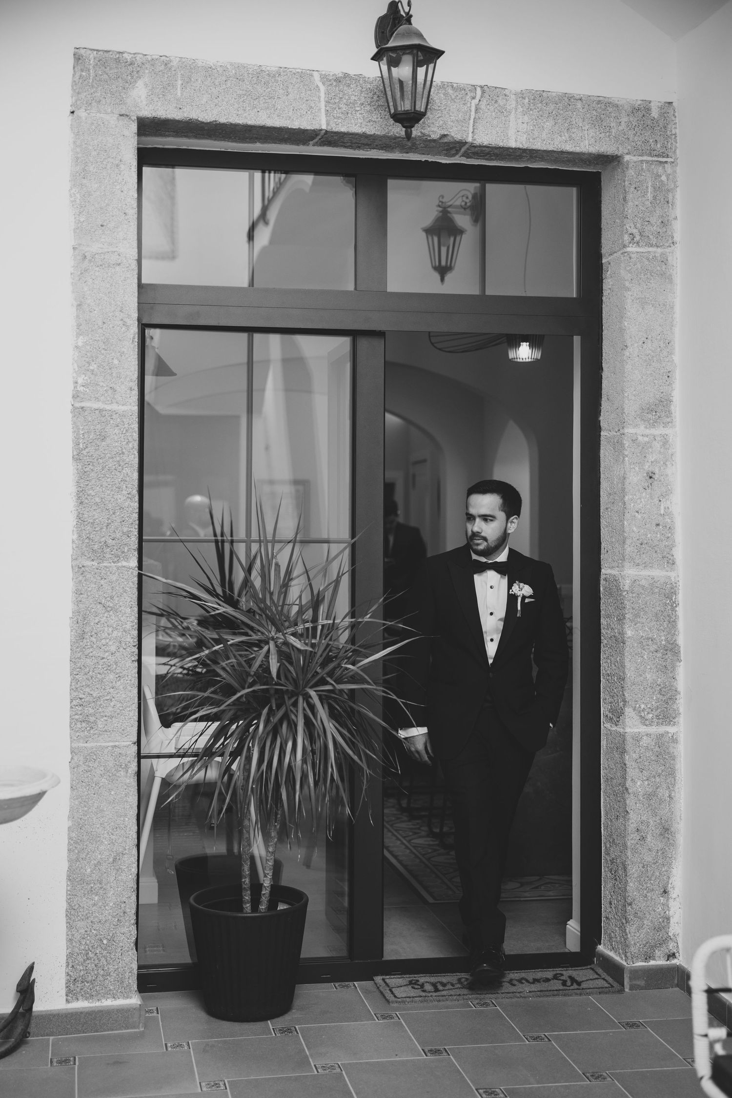Dapper groom in black tuxedo and bow tie standing in stone doorway with potted plant.