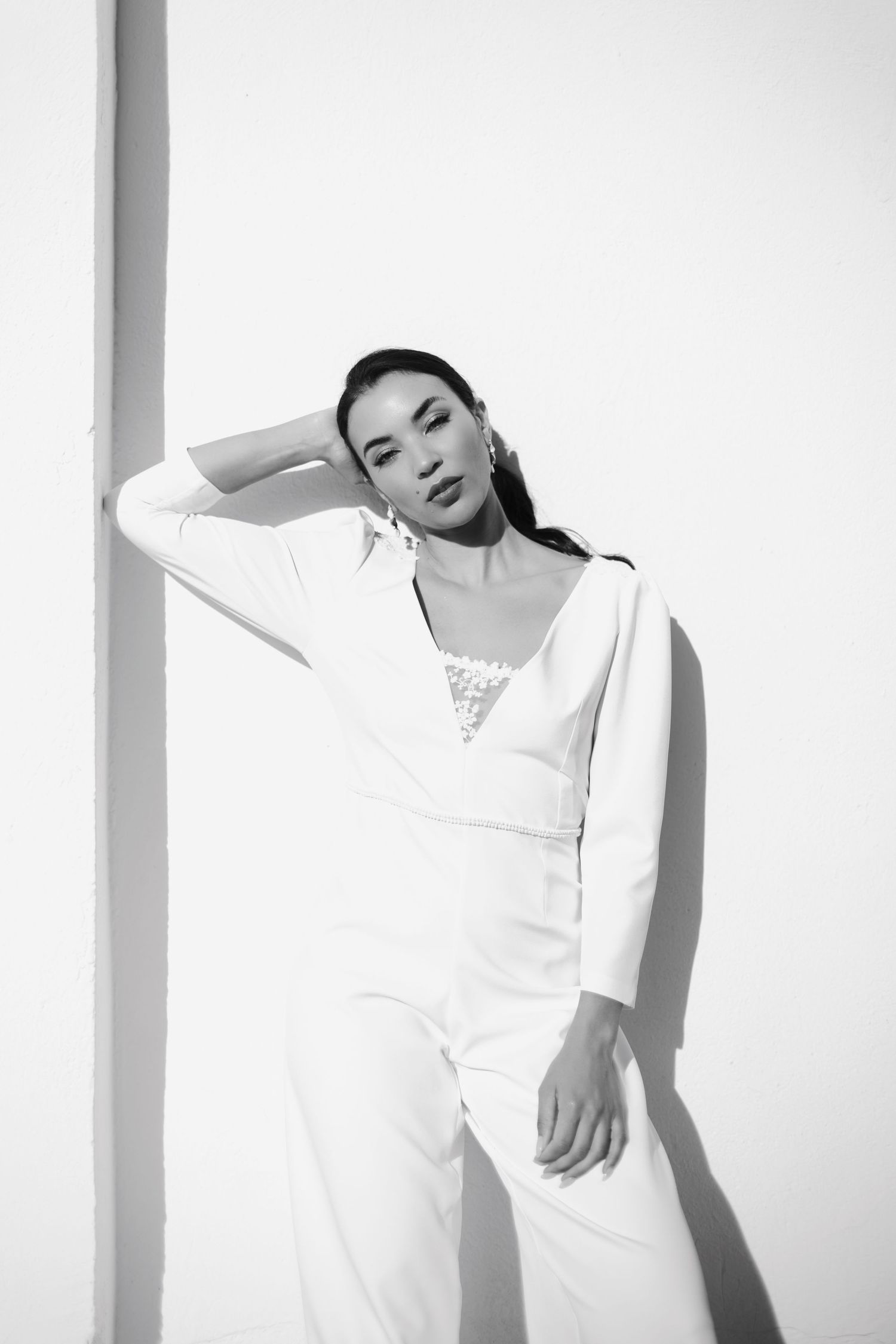 Elegant woman in white jumpsuit posing against white wall in black and white fashion photo.
