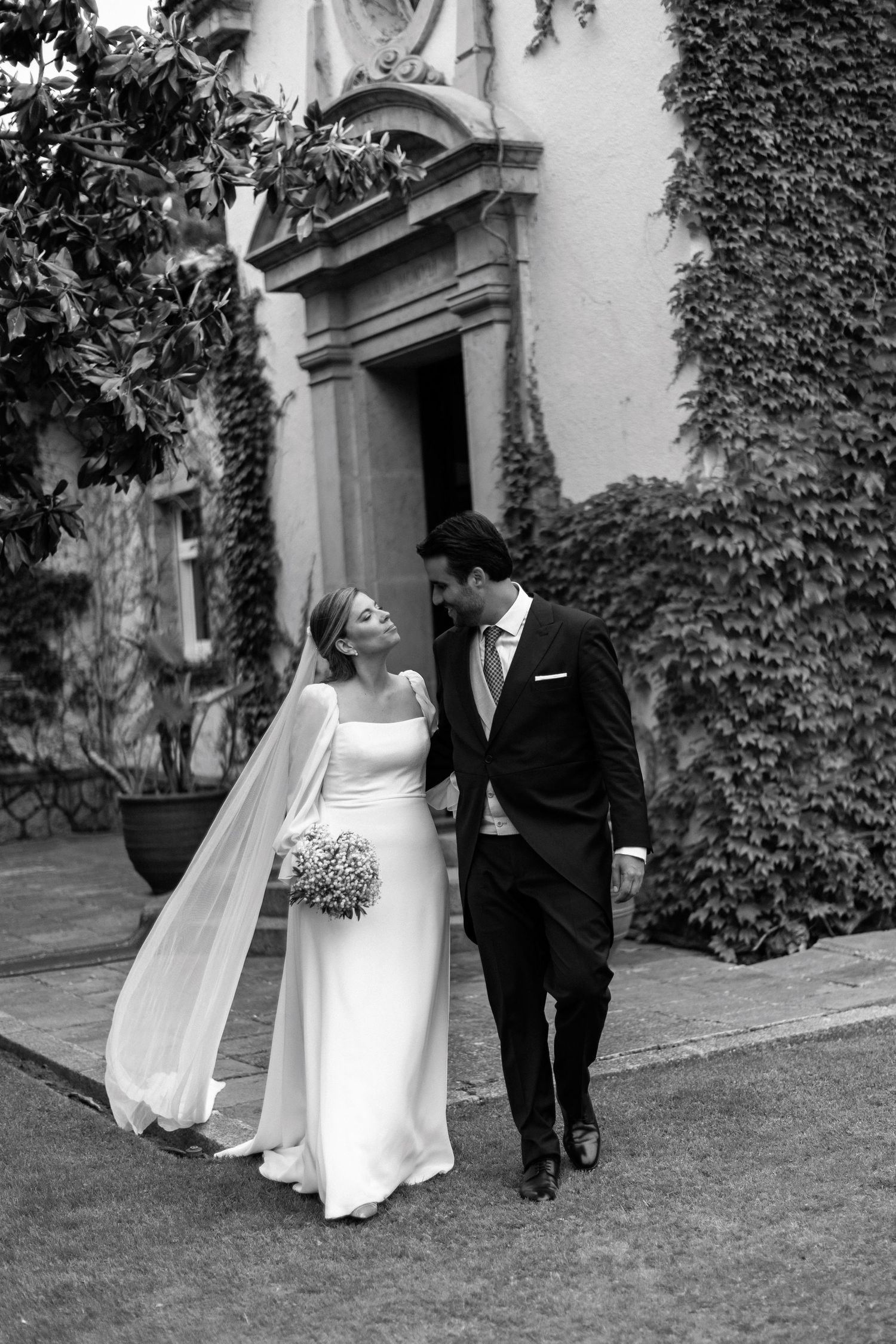 Black and white photo of bride and groom walking together outside an ivy-covered historic building.