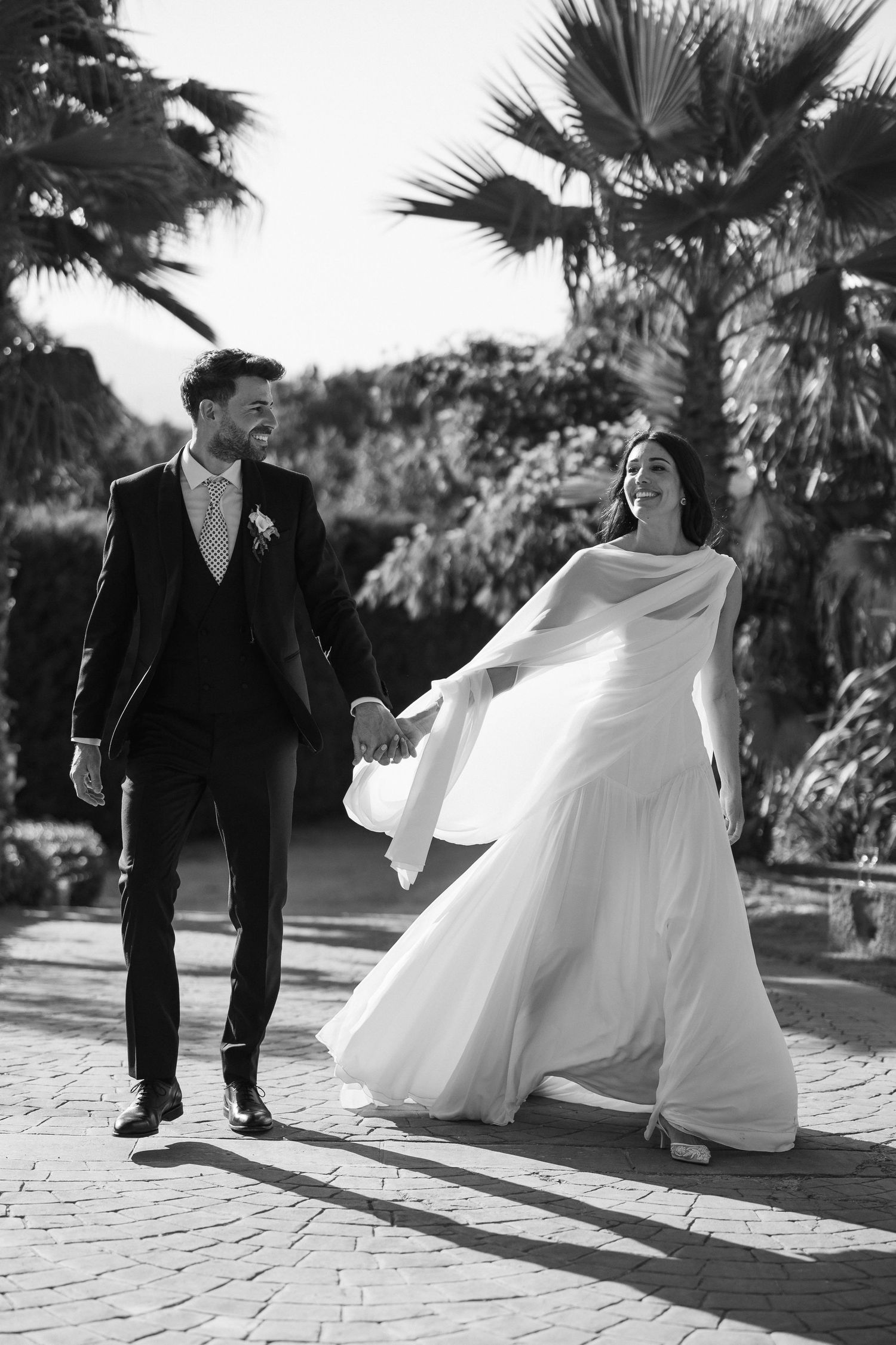Bride and groom walking hand in hand among palm trees in elegant black and white wedding photo.