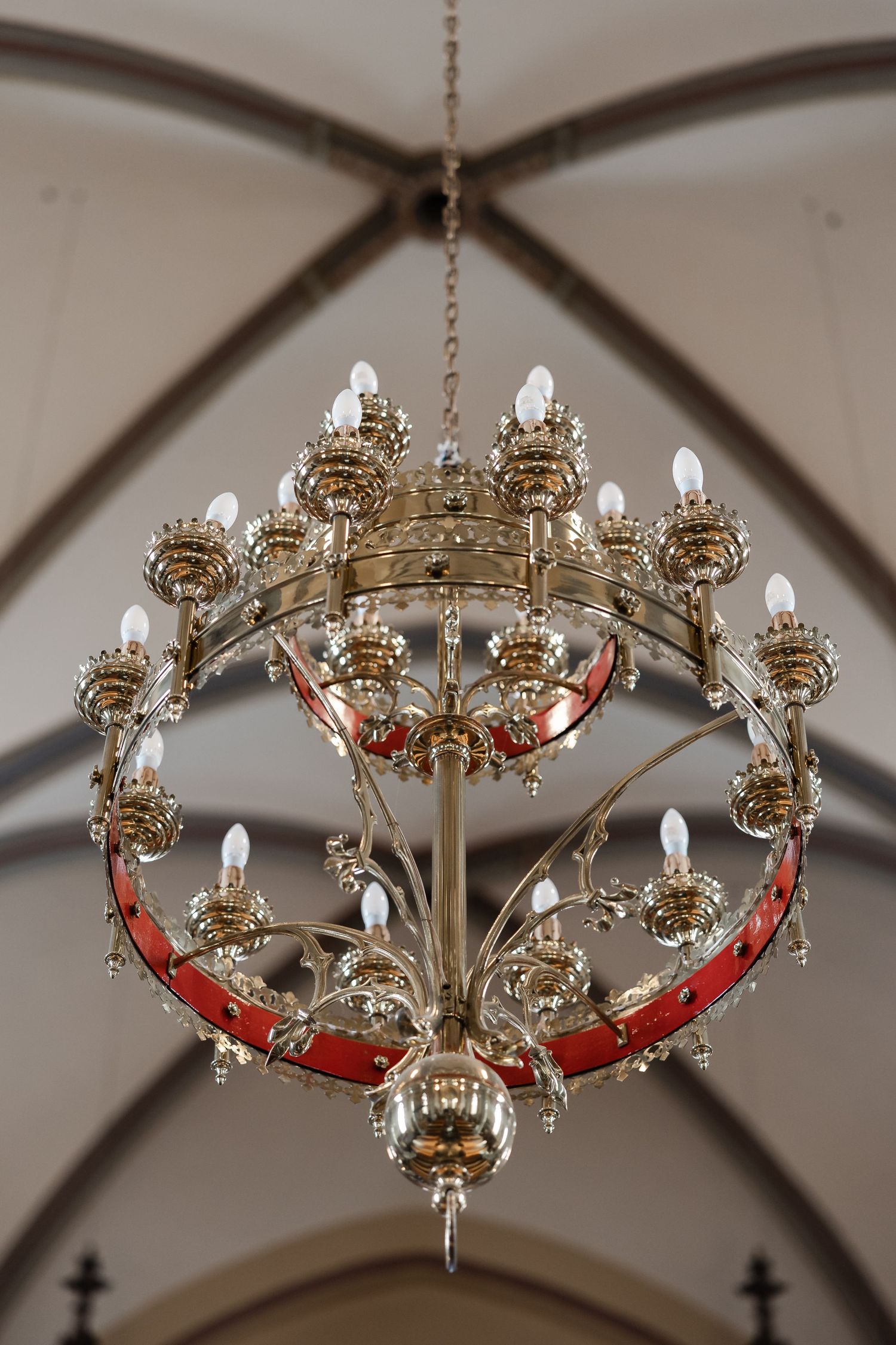 Ornate gold and red chandelier with candle-style lights hanging from a vaulted church ceiling.