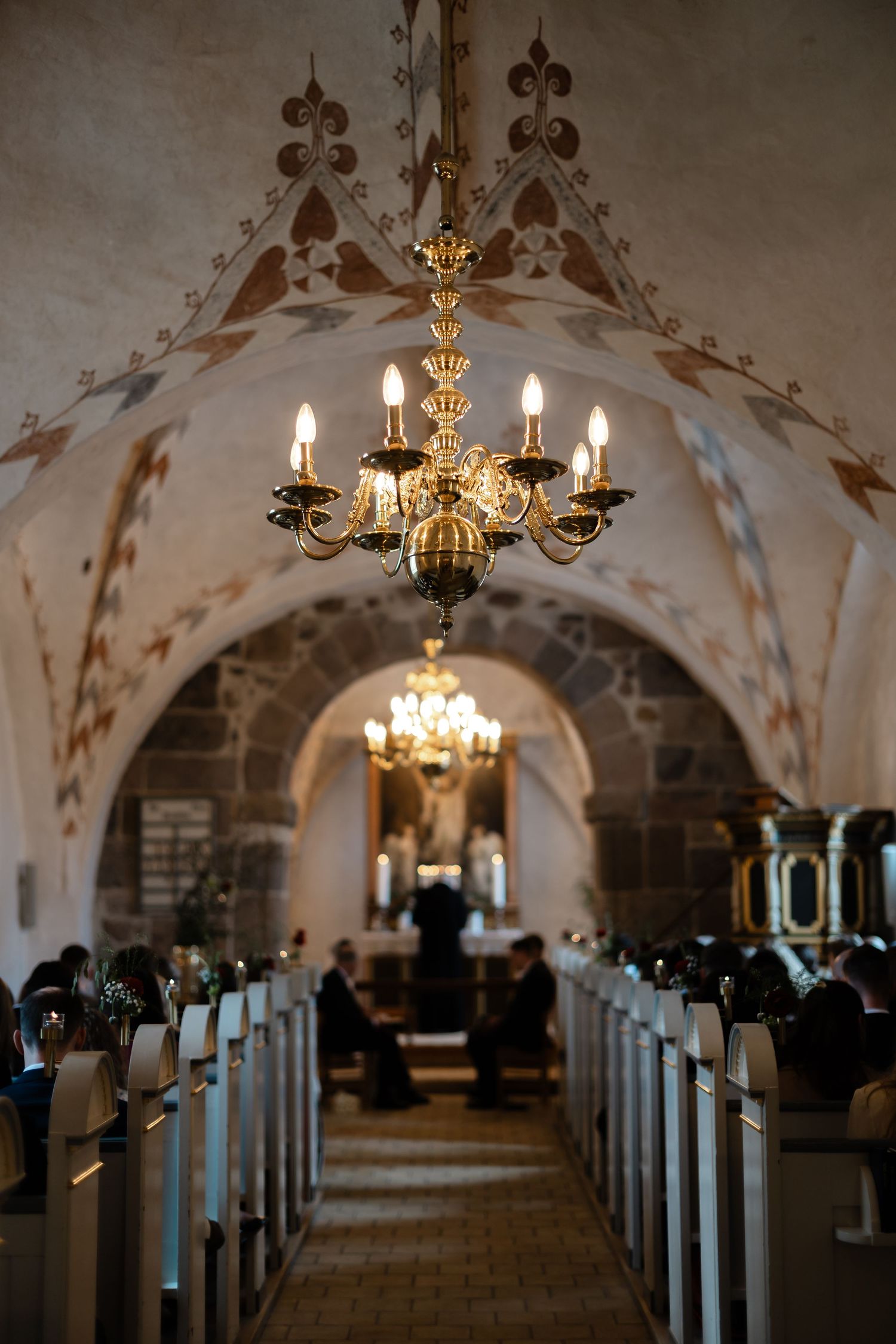 Gothic church interior with chandelier, arched ceiling murals, and pews leading to altar.