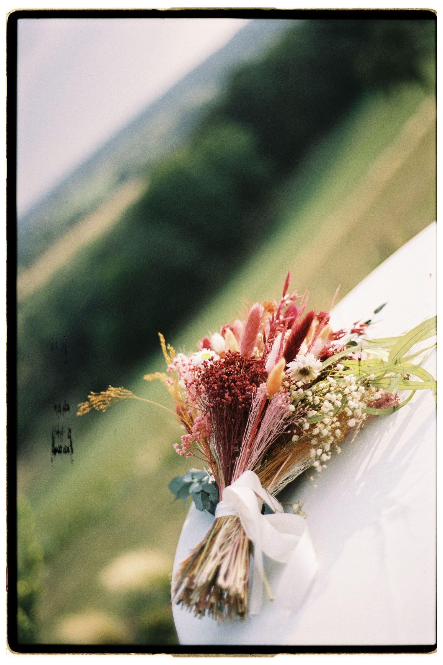 Rustic dried wildflower bridal bouquet tied with white ribbon resting on white surface with scenic backdrop.